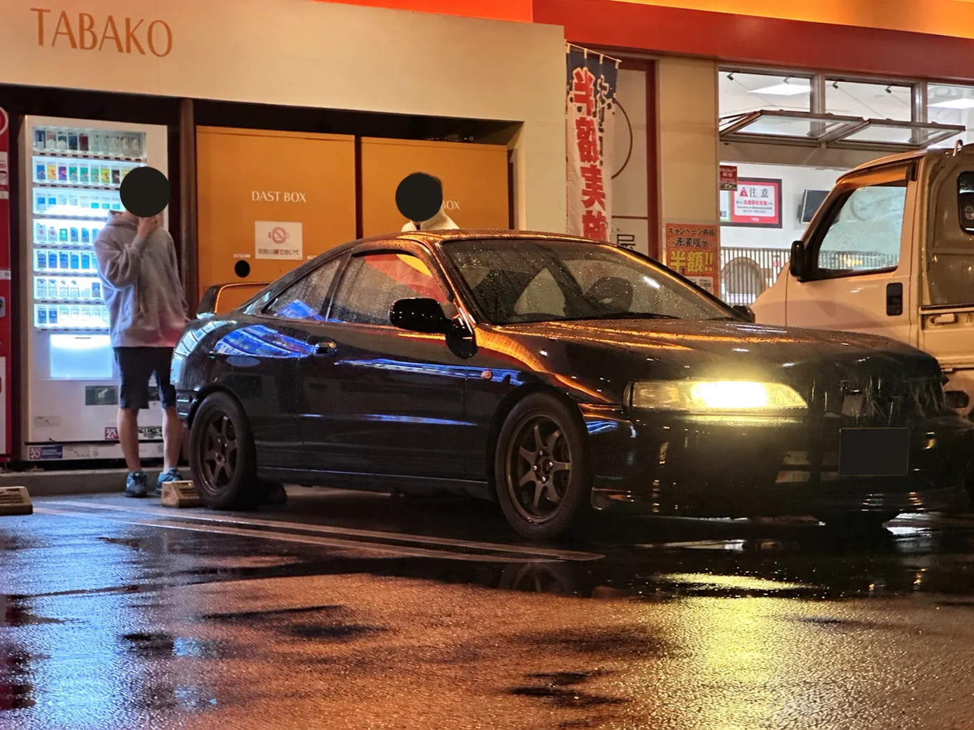 雨の夜、店舗の照明と濡れた路面に囲まれたインテグラの撮影写真をもとに、AIで顔とナンバーを匿名化した掲載用画像